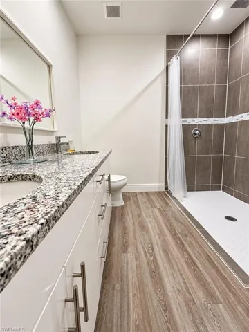 Bathroom featuring a double-sink vanity with stone-patterned countertop, white cabinetry, wood-finish flooring, and a walk-in shower with floor-to-ceiling tile surround