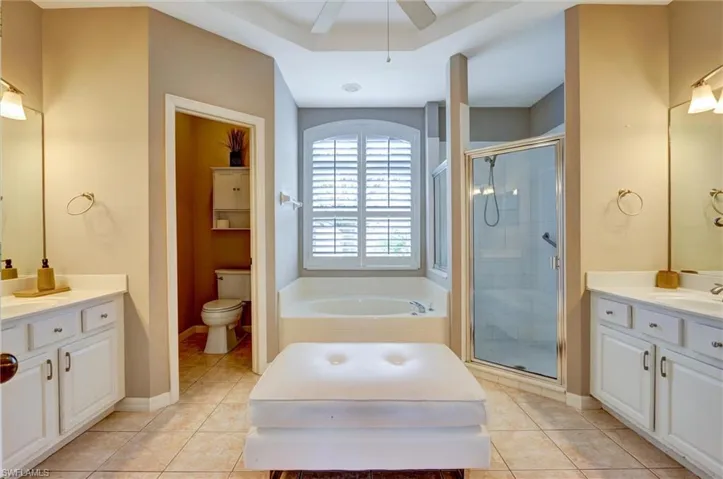 Bathroom featuring dual vanities, a built-in garden tub with arched window and plantation shutters, a glass-enclosed shower, and tile flooring