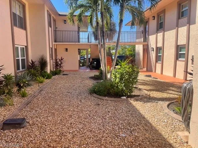 Two-story stucco exterior featuring a gravel courtyard, mature palm trees, decorative landscaping beds, and a raised metal walkway