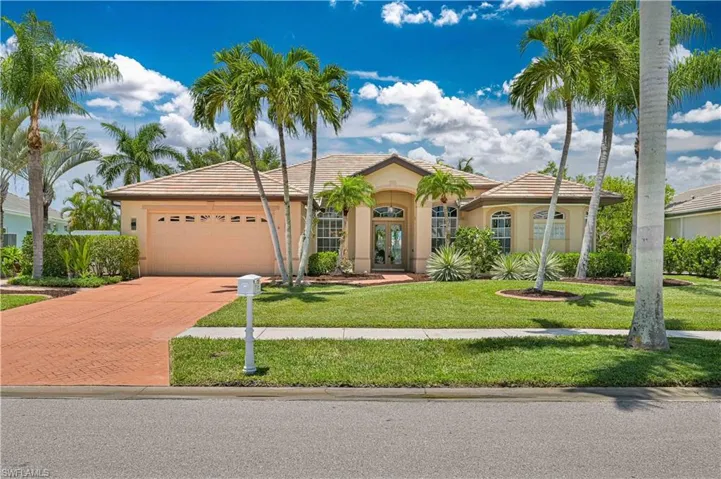 Stucco exterior with a tiled roof, featuring a brick paver driveway, an attached garage, and mature palm trees