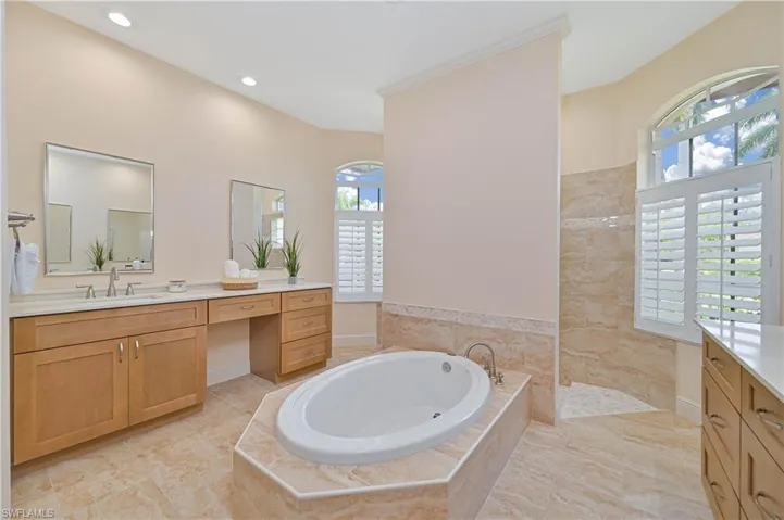 Spacious bathroom featuring a built-in soaking tub with surrounding tiled deck, dual wood-finish vanities with light countertops, arched windows with plantation shutters, a tiled shower enclosure, and recessed lighting