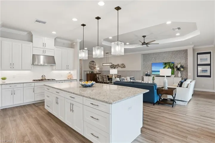 Kitchen featuring white shaker-style cabinetry, a large kitchen island with a light-toned countertop, a stainless steel range hood, pendant lighting, and wood-finish flooring extending into the adjacent living space