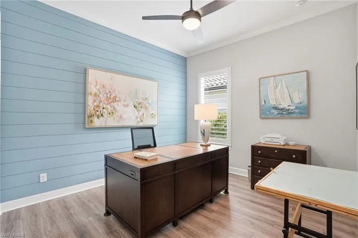 Interior room featuring a blue shiplap accent wall, wood-finish flooring, a ceiling fan with integrated lighting, a window with louvered shutters, and crown molding