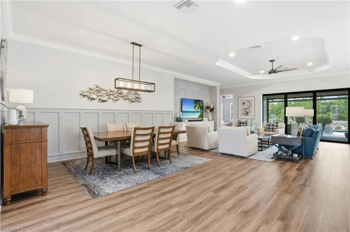 Open-concept living space featuring wood-finish flooring, a tray ceiling with recessed lighting and a ceiling fan, a dining area with board and batten wall detailing, and expansive sliding glass doors