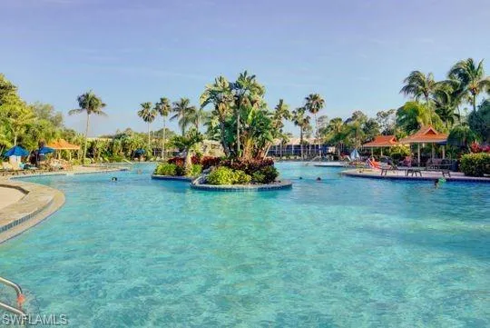 Expansive resort-style swimming pool featuring multiple sections, a central landscaped island, surrounding tropical foliage, and shaded cabanas