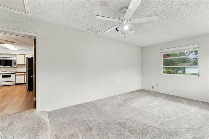 Carpeted room featuring a ceiling fan with integrated lighting, a window with horizontal blinds, and a doorway opening to a kitchen with wood-finish flooring and white cabinetry