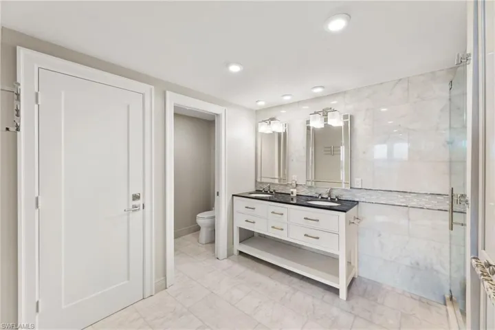 Bathroom featuring double vanity, toilet, marble finish floors, a shower stall, and baseboards