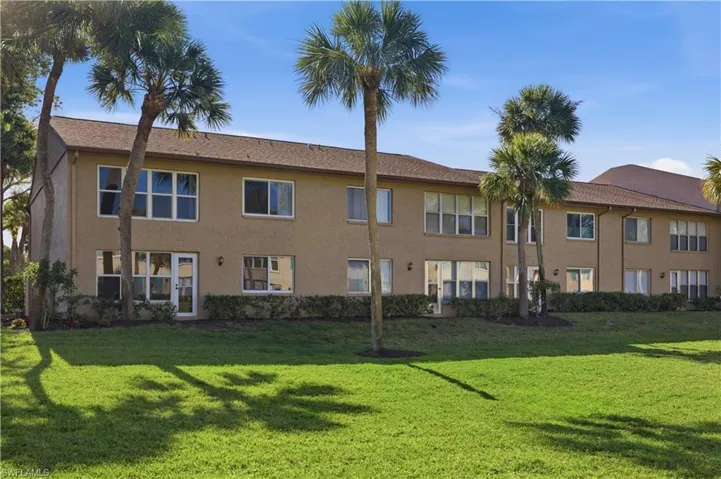 Two-story stucco building featuring multiple units with individual entrances and patios, set against a backdrop of mature palm trees and green lawn