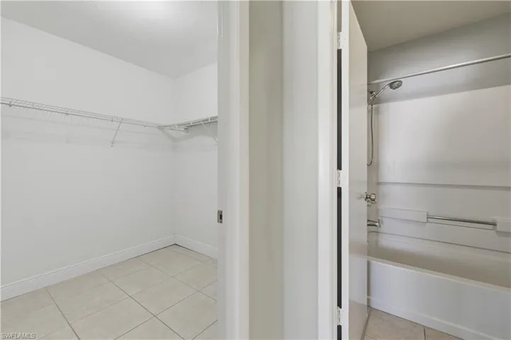 Spacious walk-in closet with wire shelving and light-toned tile flooring, positioned beside a bathroom featuring a white bathtub and shower combination with chrome fixtures