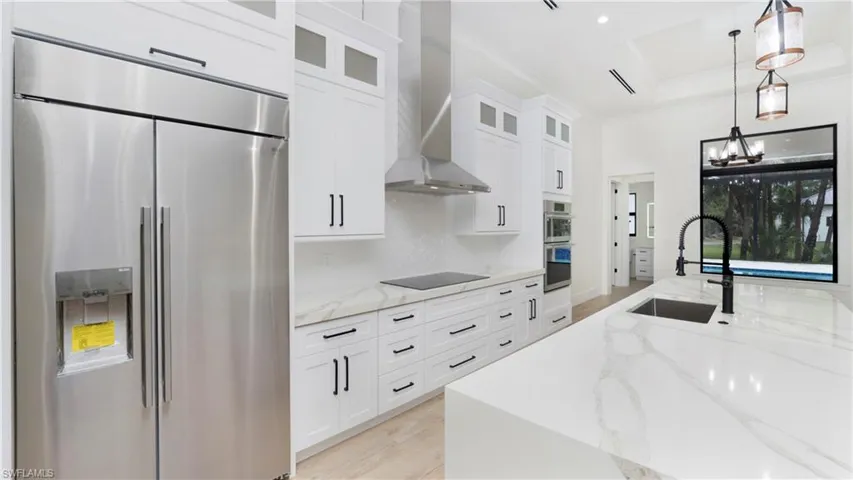 Kitchen featuring stainless steel appliances, light stone countertops, white cabinetry, wall chimney range hood, and decorative light fixtures