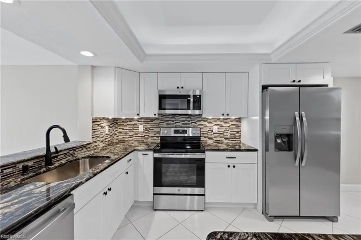 Kitchen featuring dark stone countertops, appliances with stainless steel finishes, white cabinets, light tile patterned floors, and a raised ceiling
