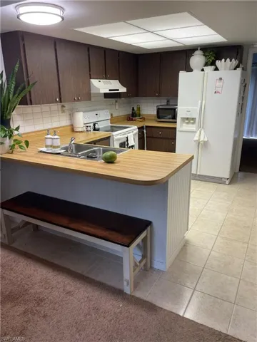 Kitchen featuring white appliances, light countertops, dark wood finish cabinetry, a peninsula, and a kitchen bar