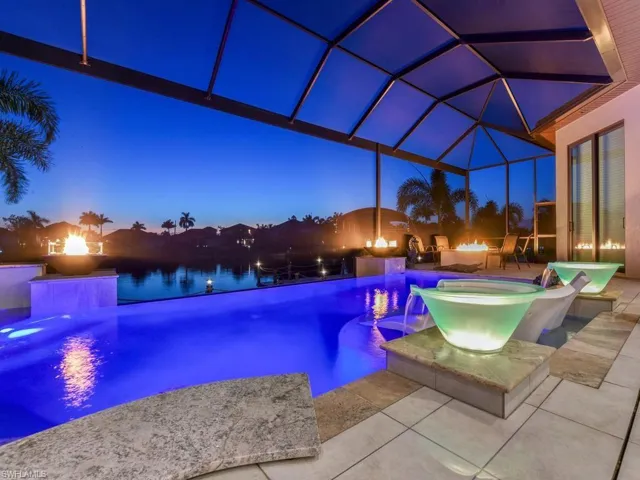 Screened outdoor living area featuring a blue-lit pool, multiple fire features, water features, and an expansive paved patio