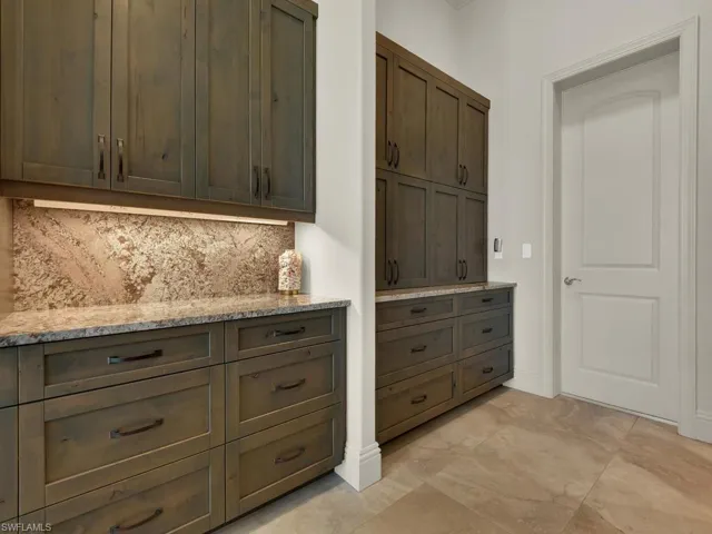 Custom wood cabinetry with dark hardware, stone countertops, integrated backsplash with under-cabinet lighting, and large format tile flooring