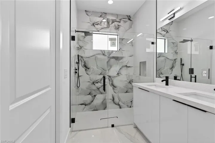 Bathroom featuring a frameless glass shower enclosure with black hardware, large format stone-look tile, a dual vanity with white cabinetry, a light-toned countertop, and matte black plumbing fixtures