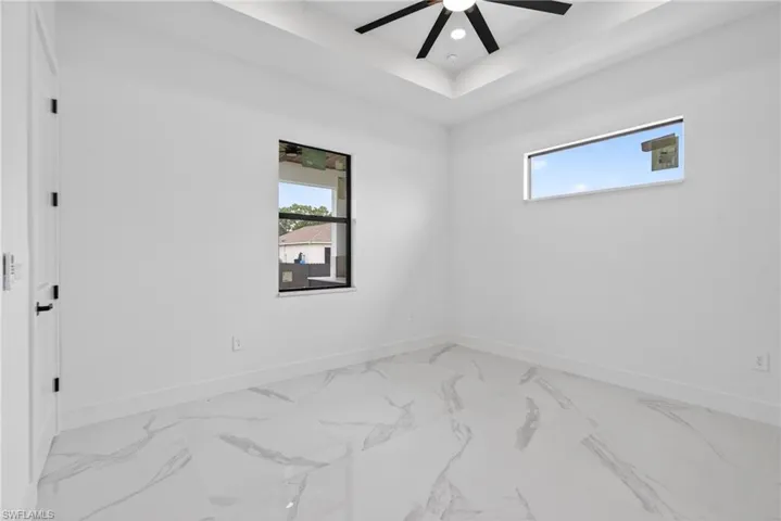 Spacious room featuring white walls, large format tile flooring, a tray ceiling with a modern ceiling fan, and two distinct window styles