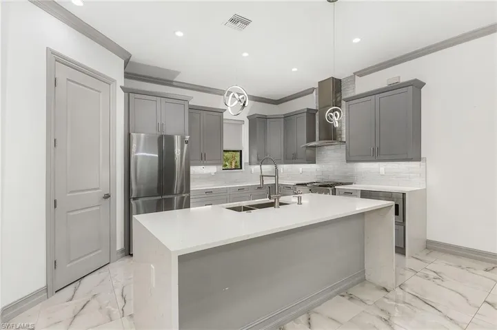 Kitchen featuring decorative light fixtures, wall chimney exhaust hood, a kitchen island with sink, and backsplash
