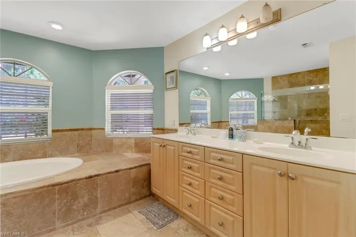 Bathroom featuring a built-in bathtub with surrounding stone tile, two arched windows with blinds, and a vanity with dual sinks