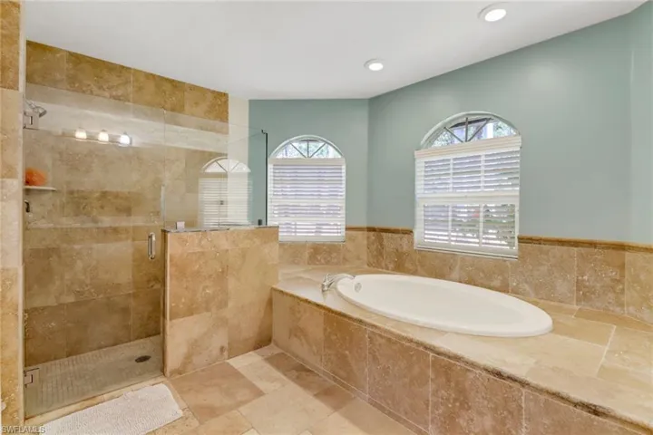 Bathroom featuring a glass-enclosed shower with stone tile, a built-in bathtub, and two arched windows with blinds