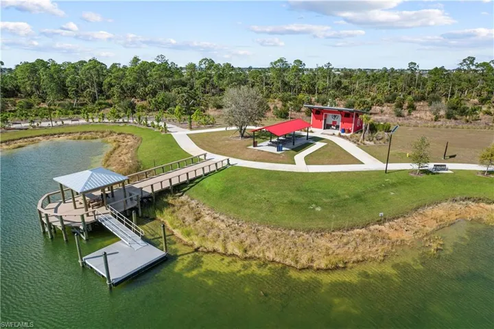 Waterfront pier with a covered seating area, floating dock, paved pathways, and a red recreational building with an attached covered picnic area
