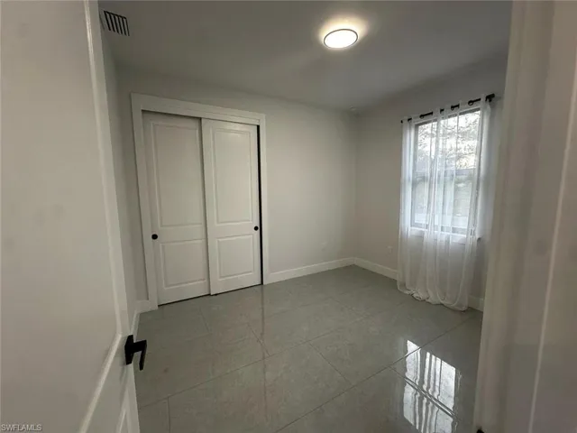 Room featuring light gray tile flooring, a window with sheer white curtains, a flush-mount ceiling light fixture, and a closet with white sliding doors and black hardware