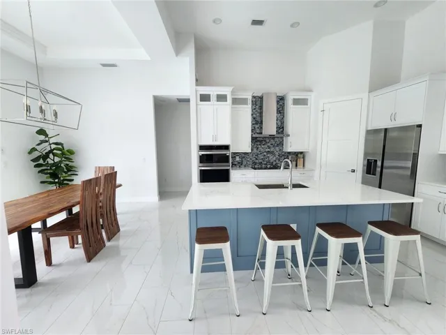 Kitchen with decorative backsplash, white cabinetry, light stone countertops, wall chimney exhaust hood, and an island with sink