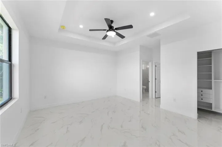 Unfurnished bedroom with a ceiling fan, multiple windows, a tray ceiling, recessed lighting, and light marble finish flooring
