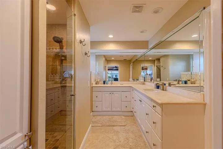 Walk-in shower with glass enclosure, dual vanity with light-colored countertops, white cabinetry with gold-toned hardware, and tile flooring
