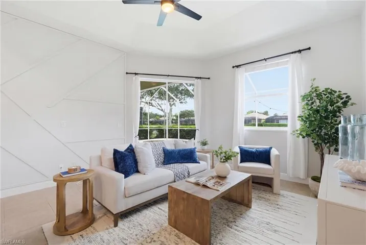 Spacious living area featuring a decorative accent wall, two large windows with white sheers, a ceiling fan with integrated lighting, and light-toned flooring