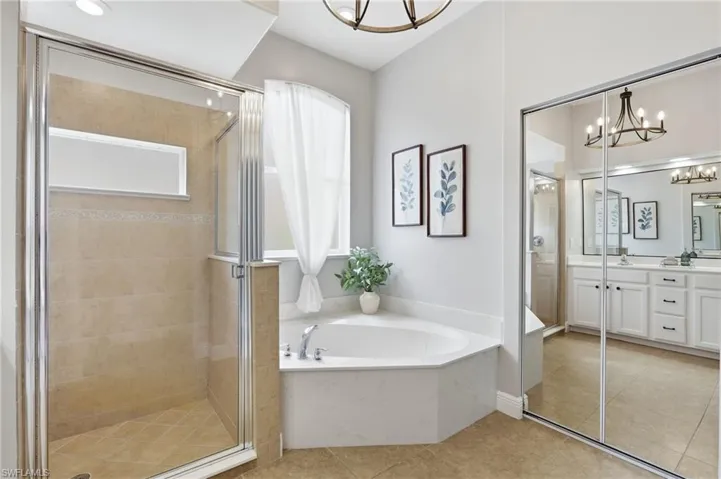 Tiled walk-in shower with glass enclosure, corner soaking tub with window, dual vanity with white cabinetry, mirrored sliding closet doors, and neutral tile flooring