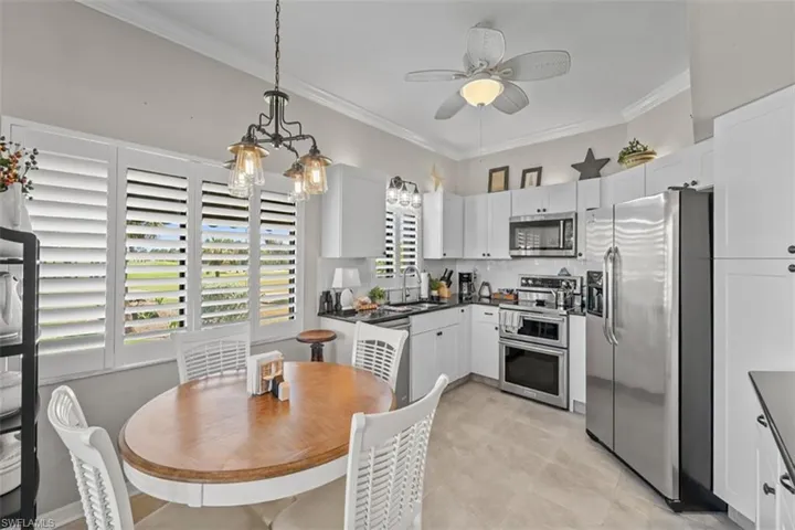 Kitchen featuring white cabinetry, stainless steel appliances, dark countertops, a ceiling fan with integrated lighting, and multiple windows with plantation shutters