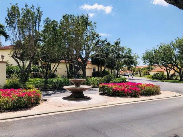 Streetscape featuring a tiered stone fountain, vibrant pink flowering shrubs, mature trees, and paved roadway