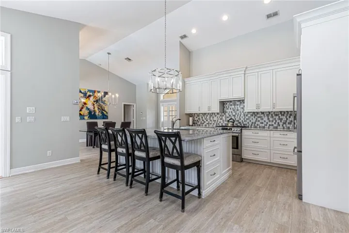 Open-concept kitchen featuring an oversized island with seating, pendant lighting, white shaker cabinetry, mosaic tile backsplash, and stainless steel appliances