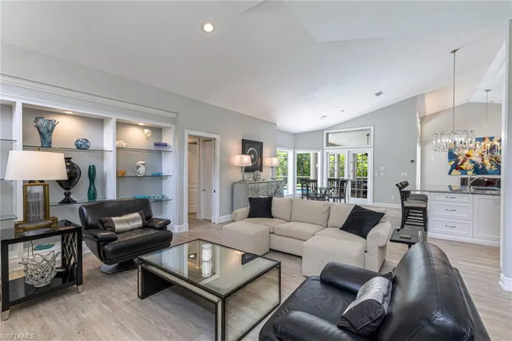 Open-concept living area featuring vaulted ceilings, light wood-finish flooring, built-in shelving with recessed lighting, a breakfast bar with stone countertop, and French doors leading to exterior