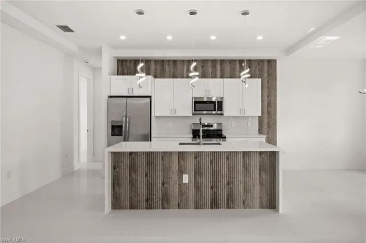 Kitchen with stainless steel appliances, light stone counters, a center island with sink, white cabinets, and hanging light fixtures