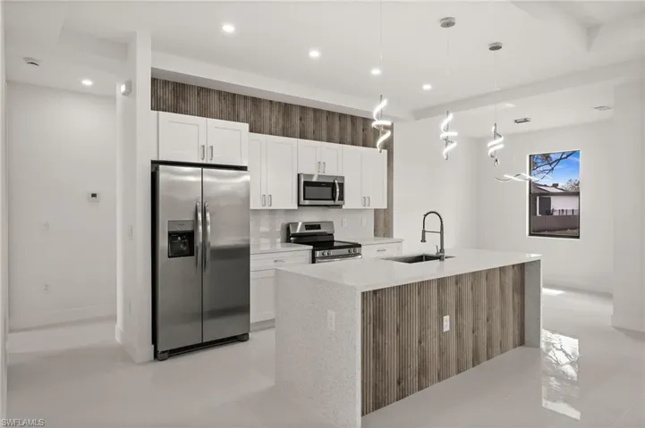 Kitchen with stainless steel appliances, white cabinets, light stone countertops, an island with sink, and hanging light fixtures