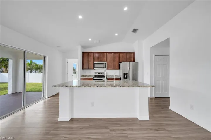 Kitchen featuring a kitchen island with sink, stainless steel appliances, light stone countertops, vaulted ceiling, and light wood-style floors