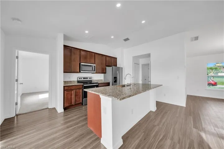 Kitchen with stainless steel appliances, a kitchen island with sink, vaulted ceiling, light stone counters, and light wood-style flooring