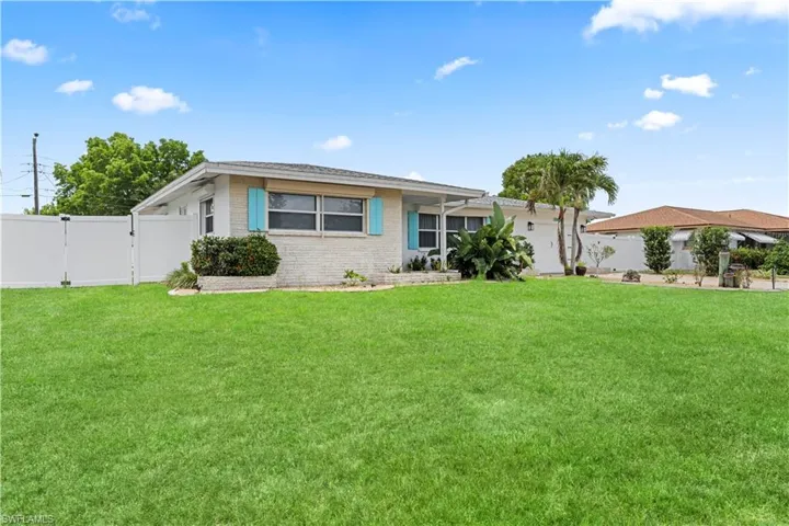 White brick exterior with turquoise shutters, a well-maintained lawn, privacy fencing, and tropical landscaping