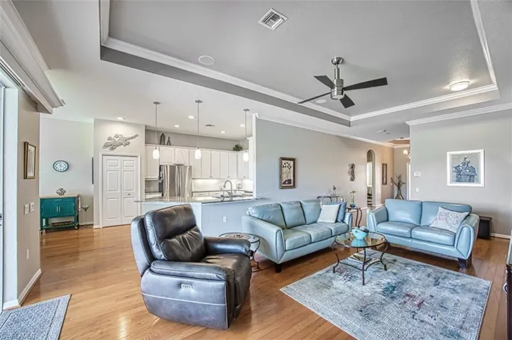 Living room with ceiling fan, a raised ceiling, ornamental molding, and light wood-type flooring