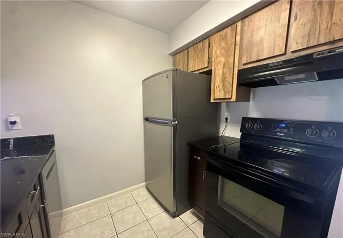 Kitchen with stainless steel appliances, under cabinet range hood, dark countertops, light tile patterned floors, and brown cabinetry