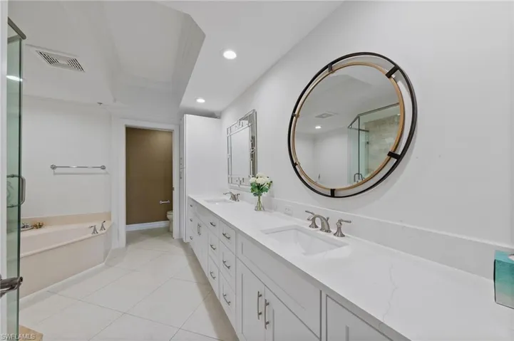 The bathroom features a double vanity with white cabinetry and countertops, a large round mirror with a dark frame, and a corner bathtub