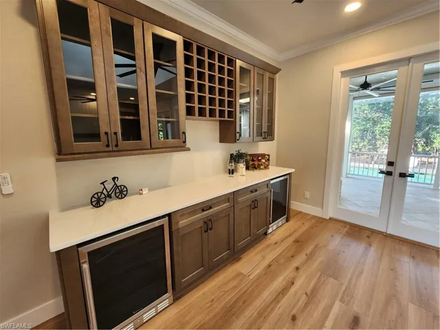 Bar area with beverage cooler, glass insert cabinets, french doors, ornamental molding, and light wood-style floors