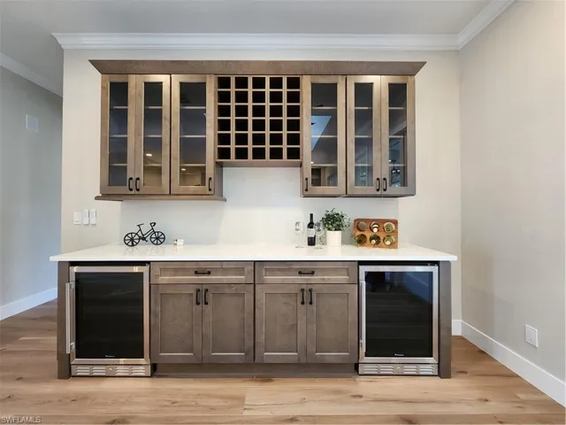 Bar with beverage cooler, glass insert cabinets, crown molding, light wood-style floors, and light stone counters