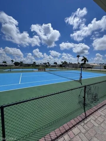 Tennis court with blue playing surfaces and green surrounds, bordered by a black chain-link fence and brick-patterned hardscaping