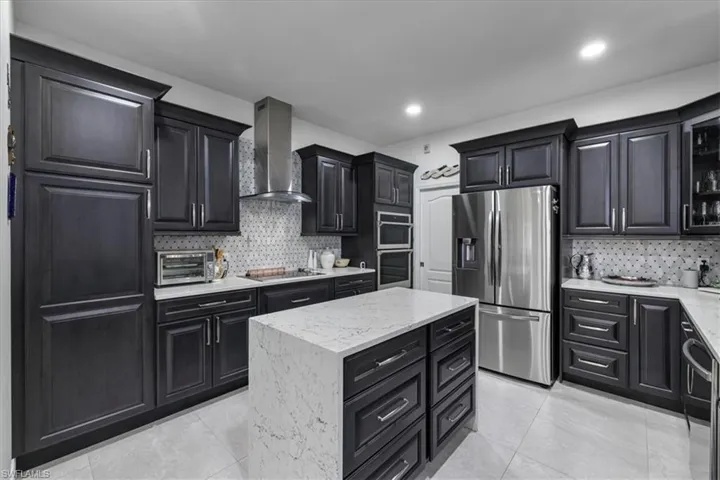 Kitchen featuring appliances with stainless steel finishes, dark cabinets, a kitchen island, wall chimney exhaust hood, and light stone counters