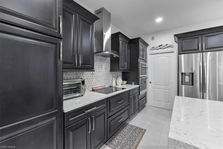 Kitchen with appliances with stainless steel finishes, light stone countertops, wall chimney range hood, decorative backsplash, and dark cabinets