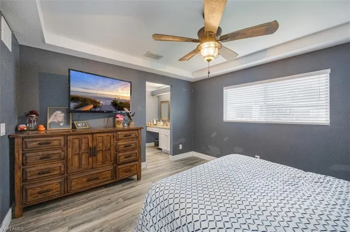 Bedroom featuring a raised ceiling, ensuite bath, light wood finished floors, and a ceiling fan
