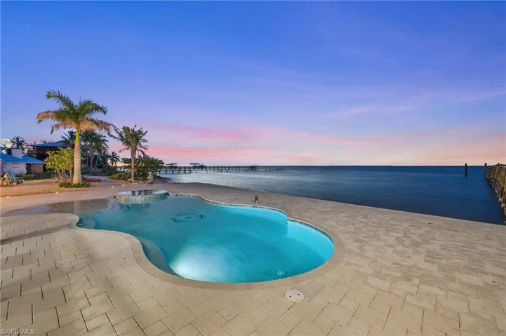Pool at dusk with connected infinity edge hot tub, a water view, and large deck with pavers.