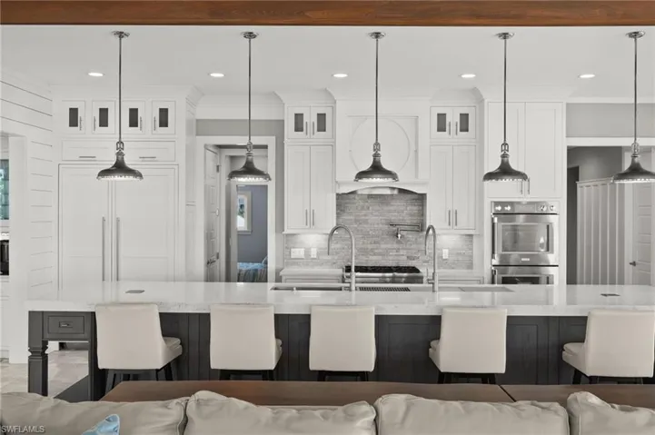 Kitchen with tasteful backsplash, stainless steel double oven, a kitchen breakfast bar, white cabinetry, a spacious island, 5 unique light fixtures and oversized island sink with sliding accessories.